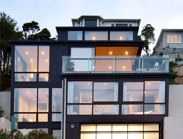 Exterior view of a contemporary multi-storey residence on a sloped site, featuring a strongly glazed facade, dark framed windows, projecting floor plates, a glass-balustraded balcony, and an illuminated garage door at ground level. The home is surrounded by landscaped planting, with another weatherboard residence visible in the background.