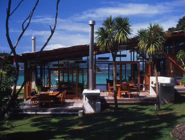 Exterior view of a low, glass-fronted coastal house opening onto a timber deck. The building has a shallow pitched roof, extensive timber-framed glazing, exposed outdoor chimneys or flues, and a patio dining area overlooking bright blue water. Palm-like plants and lawn surround the deck.