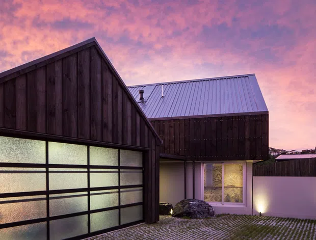 Exterior view of a compact contemporary residence at dusk, featuring dark vertical timber cladding, a steep gabled garage volume with translucent glazed garage doors, and a contrasting light metal pitched roof over the adjoining section. The foreground driveway uses permeable grid paving, with warm exterior lighting and a dramatic pink-purple sky.