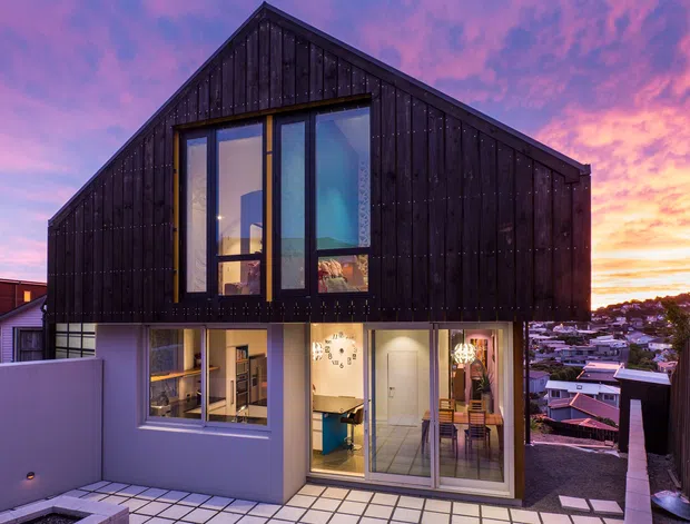 Exterior view of a compact two-storey contemporary home at dusk, featuring a steep gabled form clad in dark vertical timber boards, large floor-to-ceiling glazing, and a light-coloured lower level opening onto a paved terrace. The elevated urban hillside setting and neighbouring homes are visible beyond.