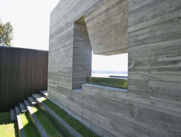 Exterior architectural view of a stark, rectilinear concrete structure with a large framed opening overlooking a distant body of water. The building has a rough horizontal board-formed concrete finish, dark vertical metal cladding, a cylindrical chimney or flue, and a series of narrow stepped grass channels and concrete-edged landscaping in the foreground.