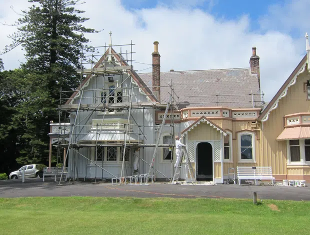 Exterior view of a grand historic timber-clad building undergoing restoration, with steep gabled roofs, ornate decorative trim, tall chimneys, verandah elements, and extensive scaffolding. The building is set beside a landscaped lawn with mature trees.