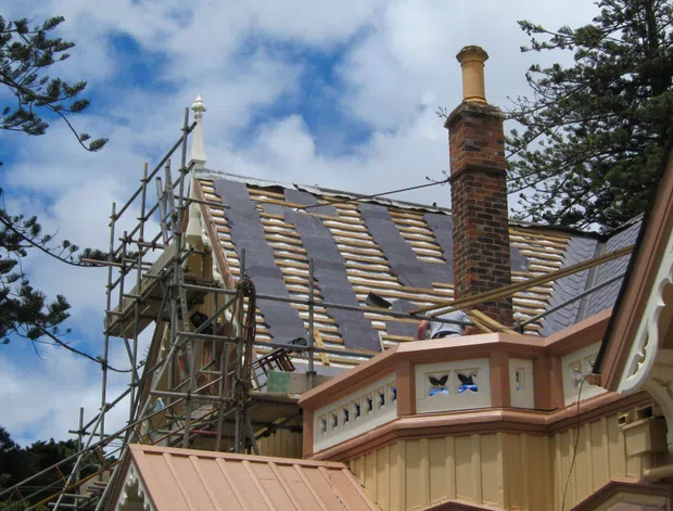 Exterior view of a heritage-style building undergoing roof restoration, with extensive scaffolding, exposed timber roof framing, slate-like roofing materials, and a tall brick chimney. Ornate painted timber trim and decorative balustrading are visible in the foreground, with mature trees and a partly cloudy sky in the background.