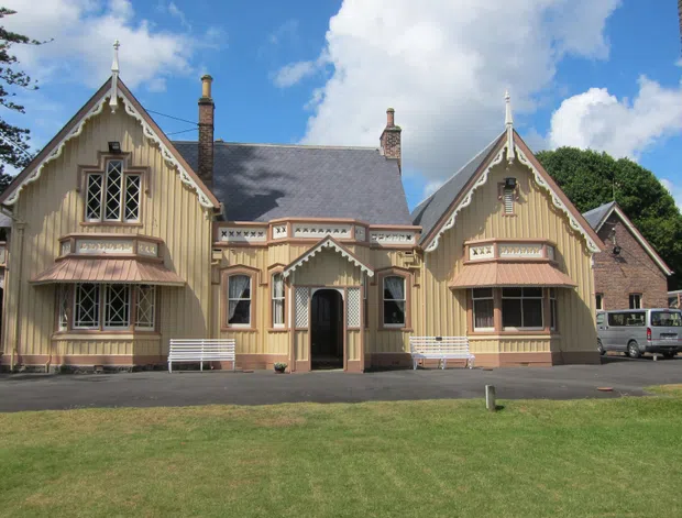 A historic timber-clad building with a symmetrical gabled facade, steep pitched roofs, ornate bargeboards, decorative window surrounds, bay windows, chimneys, and a central entrance. The building sits beside a paved forecourt and lawn, with mature trees and a bright partly cloudy sky in the background.