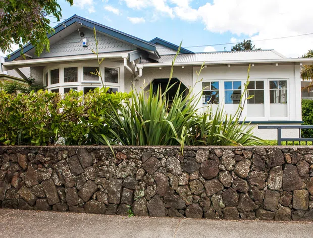 Street-facing exterior view of a single-storey bungalow-style house with a blue-trimmed gabled roof, white weatherboard walls, multi-pane windows, a covered entry porch, and a low irregular dark-stone retaining wall. Tropical planting partially screens the facade.