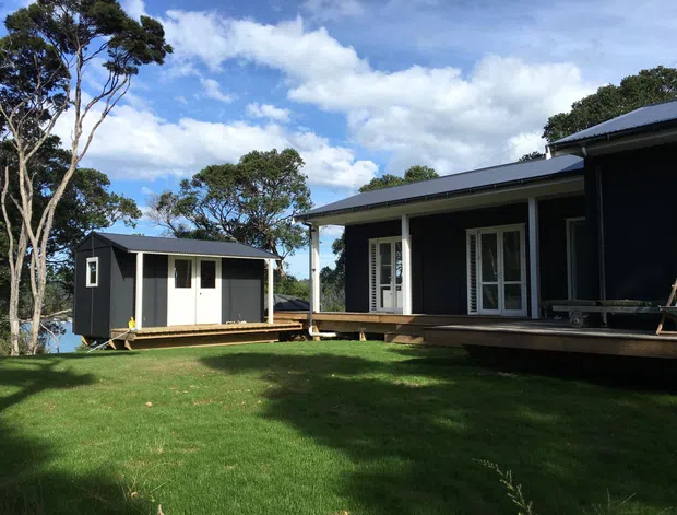 Exterior view of a dark charcoal, single-storey residential building composed of a main house and a smaller detached cabin-like structure. Both buildings have simple rectangular forms, low-pitched metal roofs, light-framed glazed doors and windows, and timber decks raised slightly above a grassy landscaped site surrounded by mature trees.
