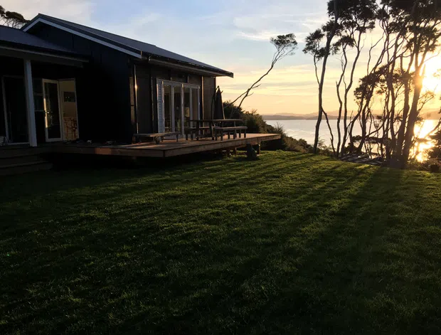 Exterior view of a dark-clad, single-storey house with a pitched roof, extensive glazed doors and windows, and a timber deck overlooking a calm coastal or lakeside setting. The building sits within a broad grassy landscape, with silhouetted trees and warm low evening sunlight.