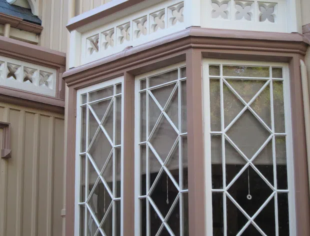 Close-up exterior view of an ornate bay window and upper decorative balustrade on a period-style building. The facade uses pale vertical cladding with contrasting dusty-rose trim, white geometric leadlight-style window framing, and pierced ornamental panels with quatrefoil motifs. The image focuses on architectural detailing rather than an interior space.