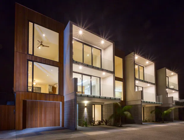 Night-time exterior view of a row of modern multi-storey attached residences. The buildings feature strong geometric forms, large illuminated glazed openings, projecting balconies, vertical timber-look cladding, pale rendered walls, and recessed exterior lighting. Garages and landscaped entry areas are visible at ground level.
