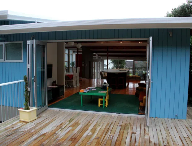 Exterior view of a compact contemporary blue-clad house opening onto a timber deck. Wide bi-fold doors reveal an open-plan interior with timber flooring, exposed ceiling beams, a dining area, living space, and a green play or recreation area. The home appears elevated, with a deck and railing visible beyond the interior.