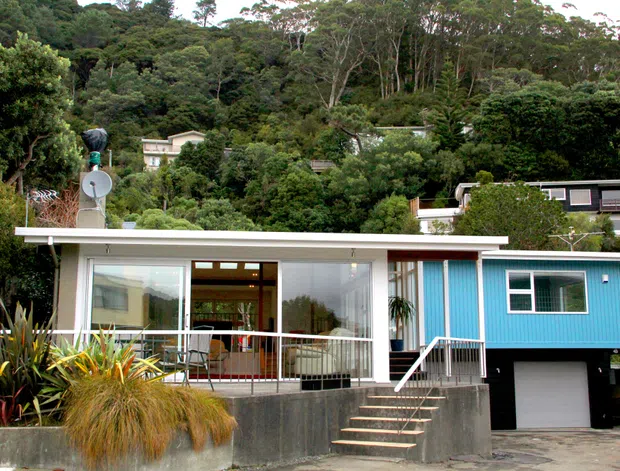 Front exterior view of a compact modern house set against a densely wooded, sloping landscape. The home has a low-pitched flat roof, white rendered horizontal volumes, extensive glazing, a vivid blue vertical-panelled upper section, an open balcony or terrace, and an integrated garage beneath the raised portion. Subtropical planting frames the concrete entry steps and retaining wall.