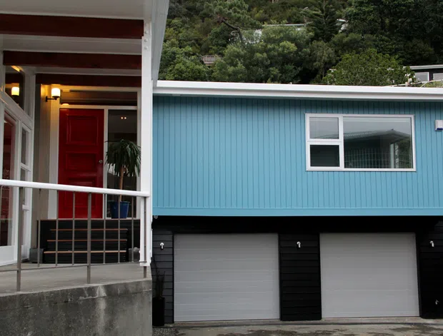 Exterior view of a contemporary residential property showing a bright blue vertical-panel upper level above a dark two-car garage. A covered entry porch with white framing, timber ceiling beams, railing, glazing, and a red front door is visible at left, with wooded vegetation behind the home.