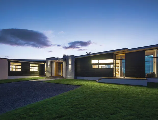Twilight exterior photograph of a low, single-storey contemporary house with strongly horizontal composition, dark cladding, broad illuminated windows, projecting roof planes, and a recessed central entry. The home sits on a lawn with a paved driveway in the foreground and an open suburban or rural setting beyond.