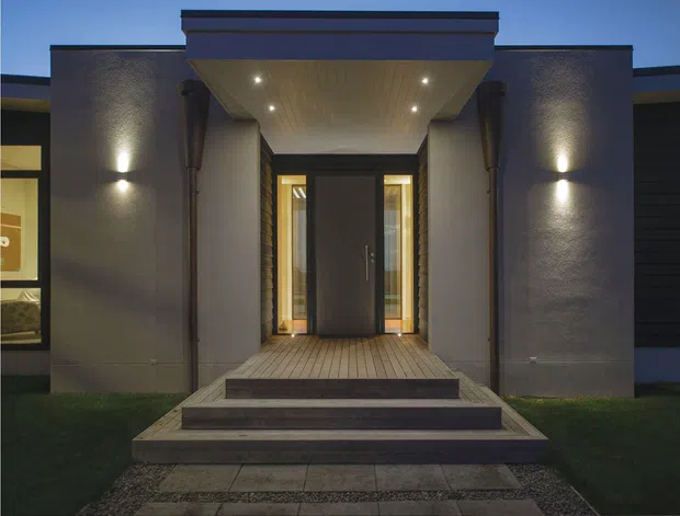 A contemporary residential entrance viewed from outside at dusk. The symmetrical facade features textured rendered walls, a projecting flat-roofed porch canopy with recessed downlights, a dark timber or metal entry door, narrow glazed sidelights, and broad timber decking steps leading to the front door. Exterior wall lighting highlights the entry surround.