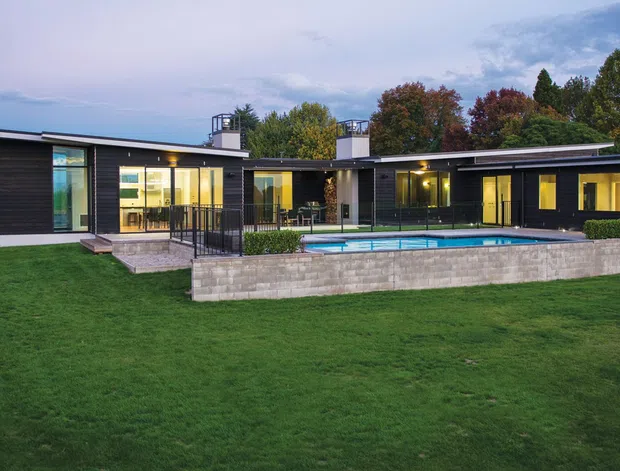 Wide exterior view of a low, single-storey contemporary house with elongated wings, dark horizontal cladding, extensive floor-to-ceiling glazing, and broad shallow roof planes. The house opens onto a manicured lawn and a raised terrace with a rectangular swimming pool, backed by mature trees. The image is captured in soft overcast or dusk-like light.