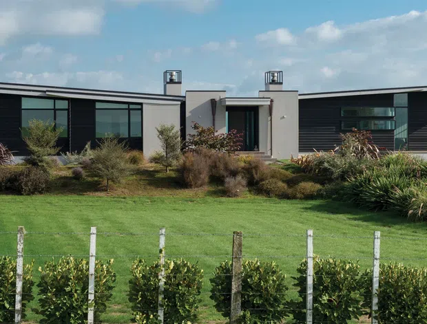 Single-storey contemporary residence viewed from the landscaped front garden, with a low asymmetrical roofline, extensive floor-to-ceiling glazing, dark horizontal cladding, and a central rendered entry volume. The property sits behind a manicured lawn and planted garden with vineyard-like rows in the foreground.