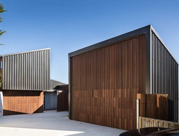 Exterior view of a contemporary multi-volume building with a sharply pitched gable roof, dark vertical corrugated metal cladding, and a prominent warm timber-clad facade. A pale concrete driveway or forecourt leads between the building volumes, with timber fencing and limited planting visible at the edges.