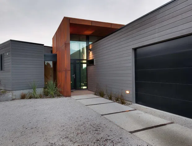 Contemporary single-storey residential facade with dark horizontal cladding, a large black garage door, and a recessed entry framed in warm timber. Concrete stepping slabs cross a light gravel forecourt, with narrow landscaping around the building.
