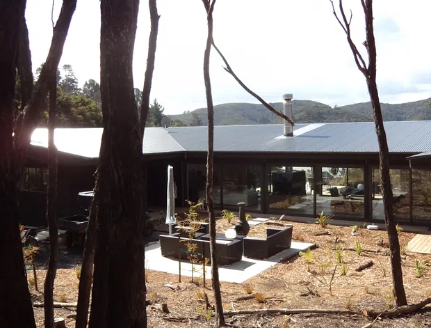 A low, single-storey contemporary house set among native bushland, viewed from the surrounding trees. The home has a broad corrugated metal roof, dark exterior walls, extensive floor-to-ceiling glazing, and a central landscaped courtyard with raised planters and a paved path. Hills and dense vegetation are visible behind the property.