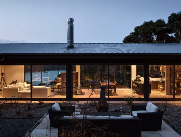 A single-storey contemporary home viewed from the landscaped outdoor area at dusk. The long, low facade has extensive floor-to-ceiling glazing, dark framing, and a broad corrugated metal pitched roof with a prominent cylindrical flue. Through the glazing, a warm open-plan living and dining area is visible, along with a kitchen at the right. The foreground contains a paved outdoor lounge with dark sofas and pale cushions, surrounded by gravel landscaping and low planting. Water is visible beyond the interior, suggesting a waterfront setting.