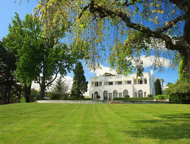 A grand white multi-storey residence set behind an expansive manicured lawn, framed by mature deciduous and evergreen trees. The facade features symmetrical composition, arched ground-floor openings, rectangular upper-level windows, and a low formal terrace wall under a bright blue sky.
