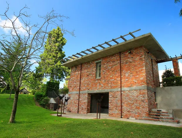 Exterior view of a two-storey brick building set within a landscaped garden. The structure has exposed red brick walls, a broad flat concrete roof slab with projecting steel pergola members, narrow vertical windows, and an open ground-level entry. A stairway and terrace connect the building to the lawn and surrounding trees.