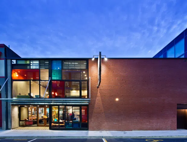 Exterior street-level view of a contemporary multi-storey brick building with extensive floor-to-ceiling glazing, vivid blue and red facade accents, and a recessed ground-level entrance. The image appears to show a commercial or institutional building at dusk, with interior activity visible through the windows and marked pavement in the foreground.