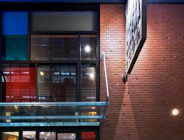 Night-time exterior photograph of a contemporary commercial building, featuring a red brick facade, large dark-framed glazed openings, a projecting illuminated sign, and a glass-balustraded entrance or balcony. Warm interior lighting and visible occupants suggest an active retail or hospitality venue.