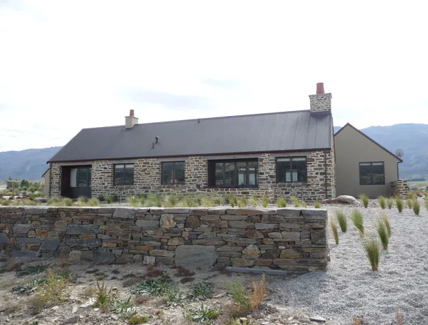 Exterior view of a single-storey rural-style stone house with a long pitched metal roof, dark-framed windows, two masonry chimneys, and a stone boundary wall. The house sits in a sparse landscaped setting with mountain terrain visible in the background.