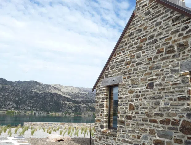 Exterior view of a small stone building with a steep gable roof, prominent masonry chimney, timber-framed openings, and a stone-paved path. The building sits beside a lake with rugged mountainous terrain visible beyond. The scene has a rustic alpine character under a bright, partly cloudy sky.