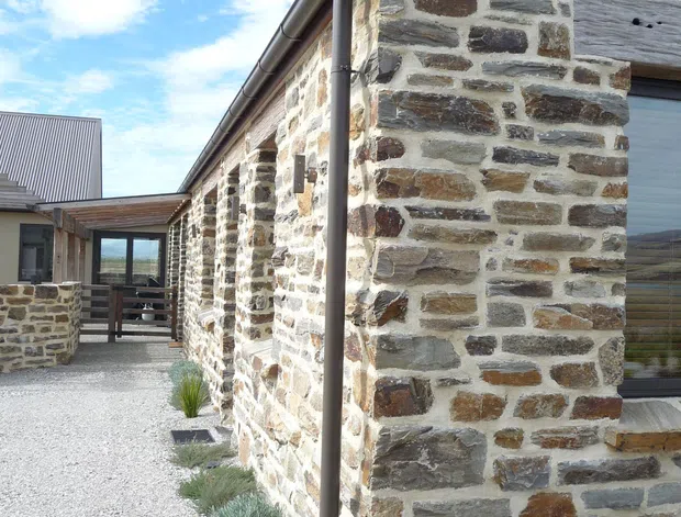 Exterior view of a low-rise building with a prominent irregular natural stone masonry facade, dark metal rainwater downpipe and guttering, and a pitched metal roof. A gravel courtyard with sparse architectural planting surrounds the building, with glazed doors and connecting wings visible in the background. The image emphasizes the stone construction and courtyard-style external circulation.
