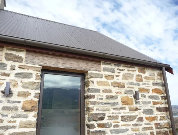 Close-up exterior view of a rustic stone building, showing irregular pale masonry walls with timber lintel detailing, a dark-framed glazed door or window, and a simple grey corrugated metal pitched roof with gutters. Mountainous terrain is faintly visible in the glazing and background.
