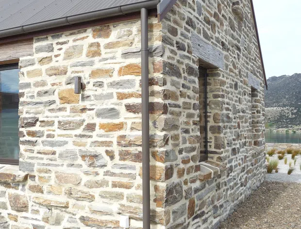 Exterior close-up of a rustic stone-clad building at a lakeside or mountainous setting. The structure has rough multicoloured masonry walls, a steep gabled corrugated metal roof, exposed stone lintels, metal gutters and downpipes, and a simple chimney. Gravel landscaping surrounds the building, with a partial view of water and rugged terrain in the background.