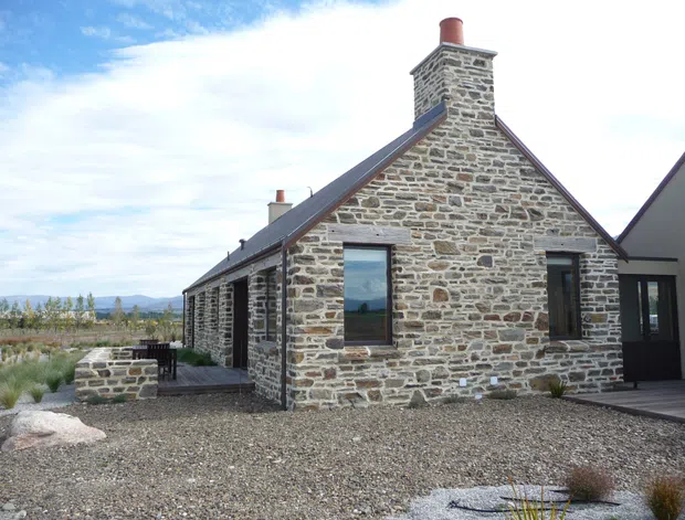 Exterior view of a low, gabled stone house with a prominent masonry chimney. The facade is constructed from irregular grey-brown stone with contrasting mortar, dark metal roof edging, and dark-framed windows and doors. A timber deck and low stone landscape wall sit beside the gravel-covered yard, with a broad rural or semi-rural landscape visible beyond.