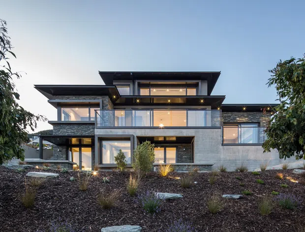A contemporary multi-level residential house viewed from the landscaped front garden at dusk. The facade combines dark horizontal roof planes, extensive floor-to-ceiling glazing, grey stone or concrete wall surfaces, and warm interior lighting. Layered projecting forms and broad overhangs create a strongly horizontal composition, with native-style planting and stone stepping elements in the foreground.