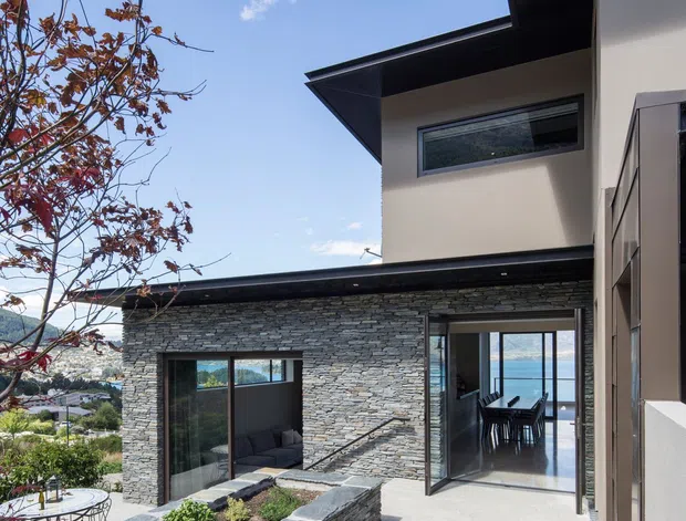 Exterior view of a contemporary multi-level home with dark projecting rooflines, pale rendered walls, extensive grey stacked-stone cladding, and large glazed openings. A landscaped courtyard with curved stone-edged garden beds and paved seating area leads to an open dining space, with a blue waterfront or bay visible through the glazing in the background.
