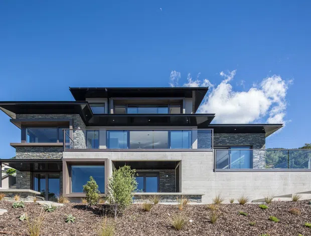 Exterior view of a large contemporary multi-level residence with strong horizontal composition, projecting flat roof planes, extensive glazing, dark stone wall cladding, and pale concrete or rendered wall surfaces. The home is set behind a landscaped frontage with sparse ornamental planting and gravel, under a clear blue sky.