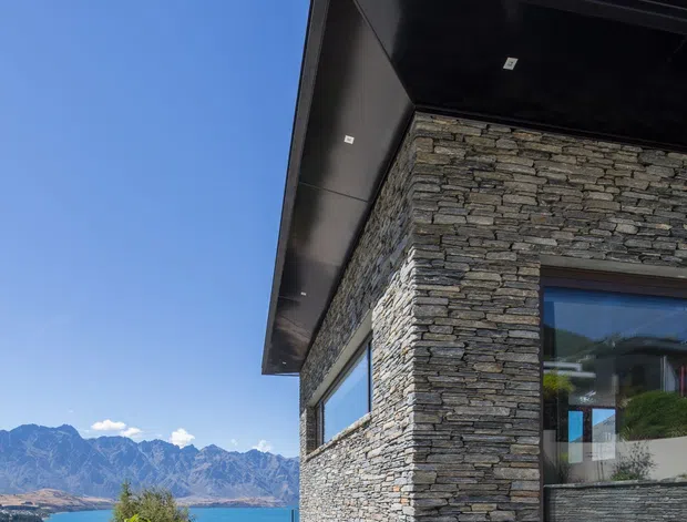 Exterior architectural photograph of a contemporary house viewed from a low angle, featuring dark stacked-stone wall cladding, expansive glazed openings, and a broad black roof overhang with recessed downlights. A landscaped garden, blue lake, and rugged mountains are visible beyond the building under a clear sky.