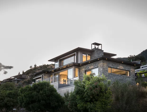 A contemporary multi-level hillside residence photographed at dusk, with extensive floor-to-ceiling glazing, projecting flat roof planes, dark metal-framed windows, and textured grey stone cladding. Warm interior lighting glows through the windows, while dense planting partially screens the lower levels and a wooded slope rises behind the home.