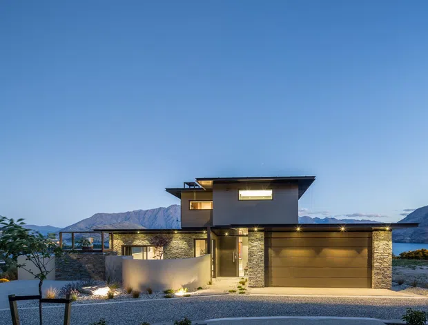 Twilight exterior photograph of a contemporary two-storey residence set against a mountainous backdrop. The house features a flat, projecting roofline, dark-framed windows, horizontal wood-toned cladding, stacked-stone wall sections, a broad integrated garage, and a softly illuminated entry. The landscaped foreground includes gravel, low planting, and a paved driveway.