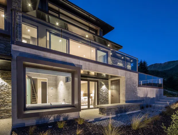 Twilight exterior view of a contemporary multi-level residence set against a mountainous landscape. The house features extensive floor-to-ceiling glazing, projecting box forms, stacked balconies, dark horizontal rooflines, stone-clad walls, rendered concrete surfaces, and illuminated interiors. A landscaped gravel garden and elevated outdoor terrace are visible in the foreground and to the right.