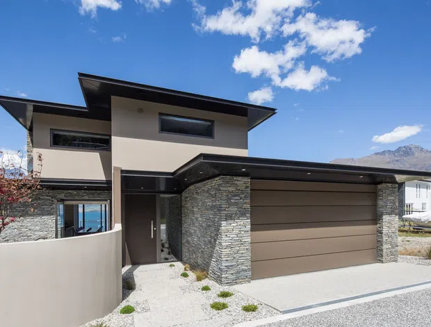 Contemporary two-storey residential home exterior with a broad low-pitched projecting roof, dark metal trim, smooth light render, stacked stone feature walls, and a double garage. The front entry is partially screened by a curved rendered wall, with gravel landscaping and mountain terrain visible in the background under a clear blue sky.