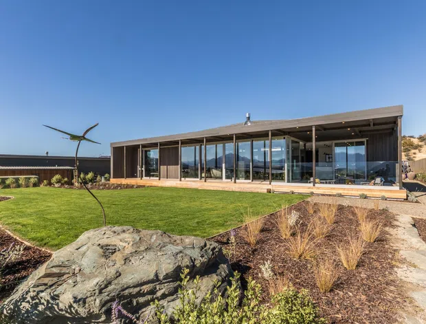 A low-profile contemporary single-storey residence set into a landscaped, rocky site. The building has a long flat roof, extensive floor-to-ceiling glazing, dark structural framing and cladding, and a broad covered outdoor edge. A manicured lawn, exposed rock outcrops, gravel paths and drought-tolerant planting surround the house under a clear blue sky.