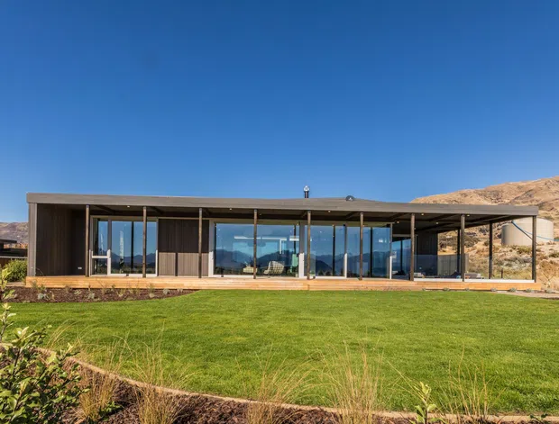 A low, single-storey contemporary residence set against a dry, rugged hillside. The home has a long, rectilinear form with extensive floor-to-ceiling glazing, dark vertical cladding, a thin flat roof, and a broad manicured lawn in the foreground. The architecture is presented from the exterior in clear daylight, with a strong indoor-outdoor connection and expansive views through the glazed facade.