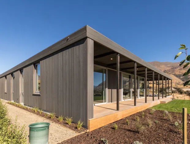 Single-storey modern house with a long, low rectangular form, dark vertical timber-look cladding, a shallow flat roof, and extensive glazed openings beneath a covered veranda. A timber deck runs along the front elevation, with slender dark posts supporting the roof overhang. The building sits in a dry, mountainous landscape with landscaped garden beds and lawn.