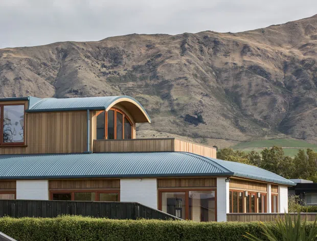 Exterior view of a contemporary low-rise home with a broad corrugated metal roof, timber-clad upper volume, curved roof element, and extensive timber-framed glazing. A rugged mountain escarpment forms the backdrop, with landscaped hedges and fencing in the foreground.