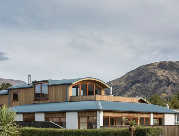 Exterior view of a contemporary multi-level mountain residence with warm vertical timber cladding, white wall panels, dark blue-grey metal roofing, and a distinctive curved upper roof and glazed balcony. The home is set against a rugged mountainous landscape, with low hedging and structured landscaping in the foreground.