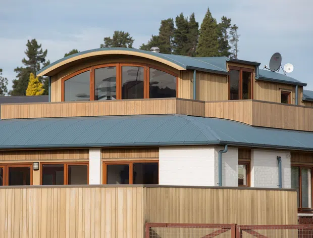 Exterior view of a multi-level contemporary residential building with curved upper-storey glazing, timber-clad balcony and privacy fencing, a blue-grey metal roof, white masonry walls, and warm timber window and door frames. Mature trees surround the property.