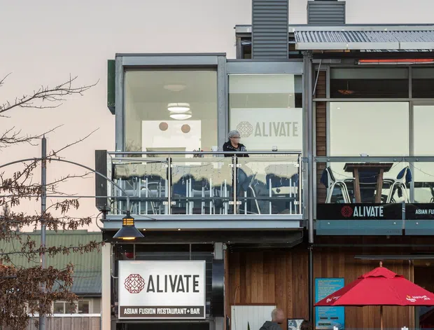 Street-facing view of a multi-storey commercial restaurant and bar facade. The building features prominent rooftop exhaust flues, corrugated metal roofing, large glazed dining areas, a glass balcony balustrade, timber-look lower facade cladding, and visible ALIVATE Asian Fusion Restaurant + Bar signage. Outdoor umbrellas and parked vehicles occupy the foreground.