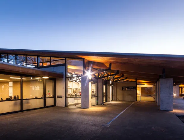 A contemporary low-rise building exterior photographed at dusk, featuring broad cantilevered flat roof planes, extensive glazed walls, exposed timber soffits, dark metal framing, and substantial concrete columns. The covered entry or circulation area opens toward a warmly lit interior and an outdoor seating area with picnic tables.
